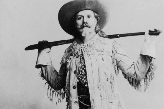 A portrait of William 'Buffalo Bill' Cody standing in front of his saddle, holding a rifle behind his back.
